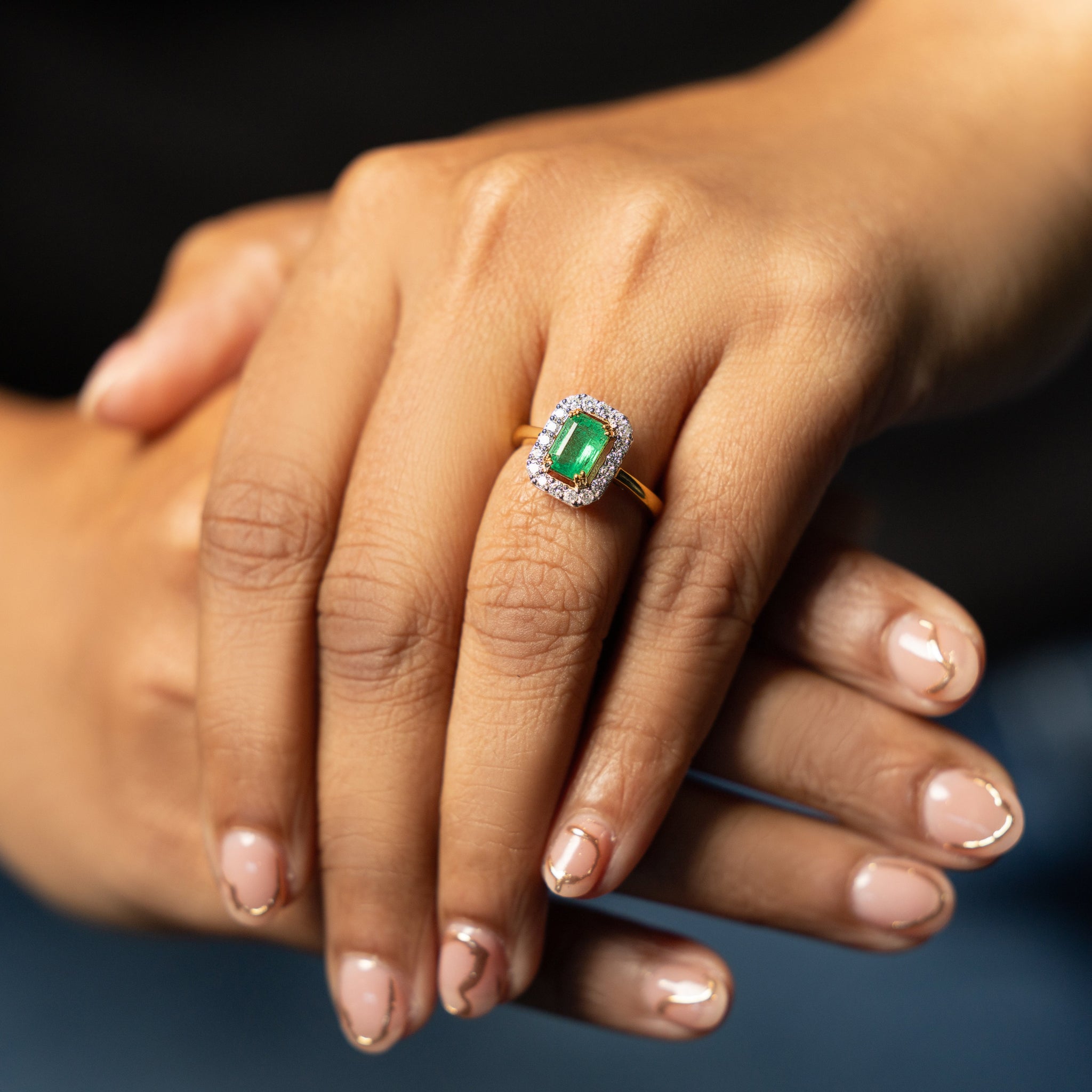 Hand wearing a ring with a green emerald gemstone and diamonds on a blurred background