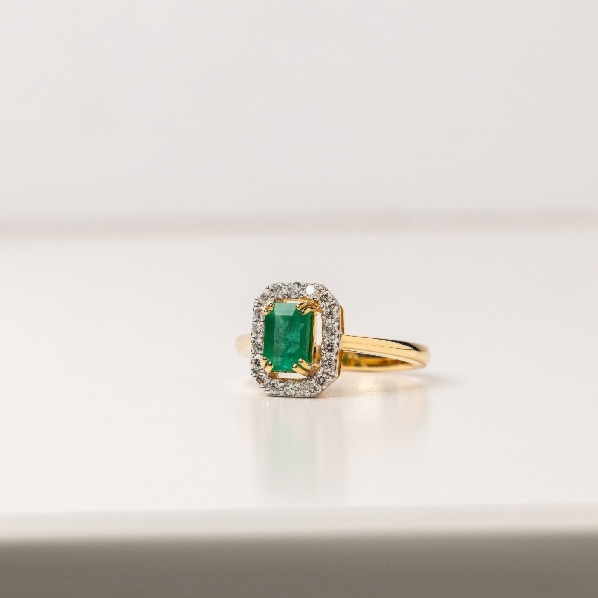 Gold ring with a green emerald gemstone and diamond halo on a neutral background